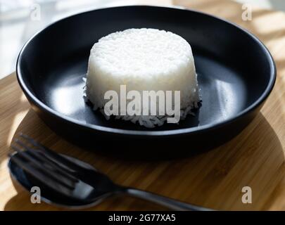 Riso di cottura a vapore di stile thailandese messo su piatto nero su sfondo di legno con coppia di forchetta posate. Foto Stock