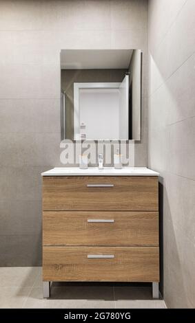 Bagno moderno e rinnovato in stile minimalista con pareti piastrellate grigie, specchio e cassetti in legno sotto il lavabo. Foto Stock