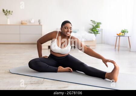 Donna nera curvy che allunga le gambe, si esercita sul tappeto di yoga, leader di stile di vita attivo, cercando di perdere peso a casa Foto Stock