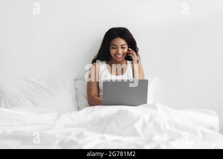 Sempre in contatto. Giovane afroamericana seduta a letto, lavorando su computer portatile e parlando sul cellulare Foto Stock