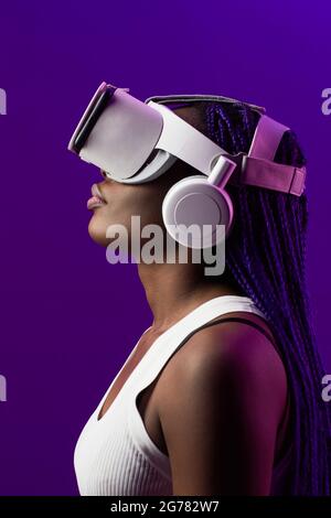 Ritratto futuristico con vista laterale di una giovane donna afroamericana che indossa un visore VR su sfondo viola Foto Stock