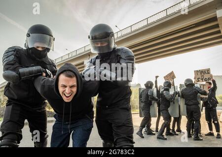 Ufficiale di rivolta in caschi torcendo le braccia del protettore aggressivo in felpa con cappuccio mentre lo arrestano sulla strada Foto Stock