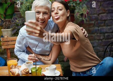 Una giovane ragazza sta facendo i volti divertenti mentre prende un selfie con la sua nonna in un'atmosfera piacevole ad un bar. Tempo libero, bar, amicizia, all'aperto Foto Stock