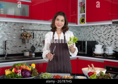 Bella donna asiatica di mezza età casalinga che indossa grembiule in piedi nuova cucina tono rosso e tenendo verdure fresche verdi e coltello di preparazione di verdure Foto Stock