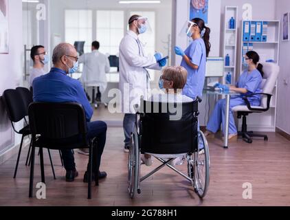 Medico specialista con visiera e maschera di protezione che parla con assistente in piedi in sala d'attesa della clinica medica prima dell'esame dei pazienti. Persone che visitano Medic durante la pandemia globale del covid 19 Foto Stock