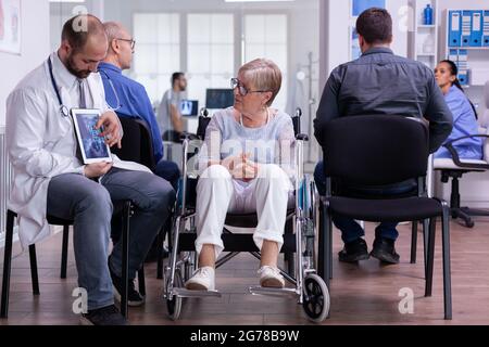 Medico uomo seduto nell'area dell'ospedale di attesa con la donna anziana di disabilità che discute di trattamento di recupero contro la malattia di mezza età. Medico che mostra la radiografia delle ossa utilizzando un computer tablet Foto Stock