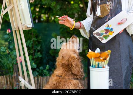 Giovane artista femminile che lavora sulla sua tela d'arte dipingendo all'aperto nel suo giardino con Golden Retriever mantenendo la sua azienda. Concetto creativo di hobby. Foto Stock