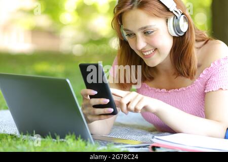 Felice studente che indossa le cuffie utilizzando il computer portatile e lo smartphone in un parco Foto Stock