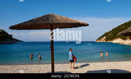 Isole IONIE, Ithaca, isola di Odysseus, vicino a Vathi, Filiatro Beach, spiaggia di ghiaia fine, la gente sulla spiaggia, ombrellone rotondo coperto di paglia di bambù, la gente nell'acqua, cielo blu con le nuvole, una singola barca a vela è ancorata nella baia Foto Stock