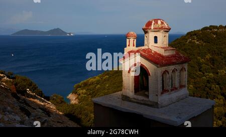 Isole IONIE, Ithaca, isola di Ulisse, costa nord-orientale, Marmakas Bay, L'isola di Atokos si può vedere sullo sfondo a sinistra, una cappella in miniatura sulla strada a destra, il mare è blu scuro, il cielo è blu chiaro Foto Stock