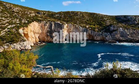 Isole IONIE, Zante, Costa occidentale, Mare Adriatico, spiaggia, porto, Porto Limnionas, vista da un po 'sopra la baia, cespugli verdi in primo piano, mare con spray surf bianco a metà distanza, acqua verde-blu, roccia sovracresciuta faccia opposta, cielo blu con una singola nuvola bianca Foto Stock