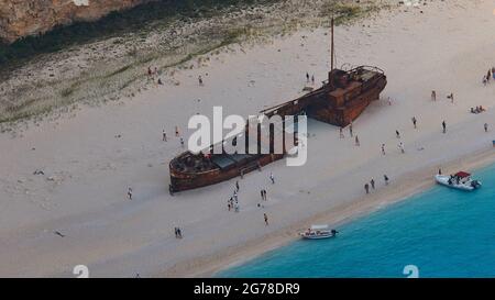 Zakynthos, Paralia Navagio, Shipwreck Beach, vista dall'alto del relitto della MV Panagiotis che giace sulla spiaggia, persone nel cielo intorno al relitto, telefoto, triangolo in basso a destra con mare verde-blu, barche da escursione all'ancora sulla spiaggia Foto Stock