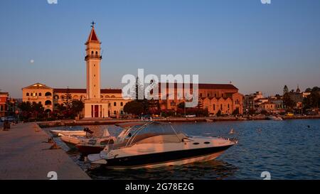 Città di Zante, luce del mattino, Chiesa di San Dionisios, barche nel porto in primo piano, cielo blu, luna accanto alla torre della chiesa Foto Stock