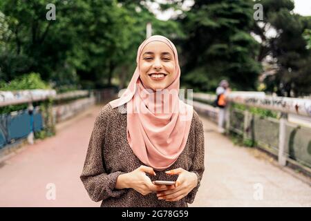 Bella ragazza musulmana che cammina nel parco e digitando nel suo telefono cellulare. Sorride e guarda la macchina fotografica. Foto Stock