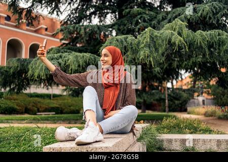 Bella donna musulmana che indossa hijab seduto nel parco e prendendo un selfie con il suo telefono. Foto Stock