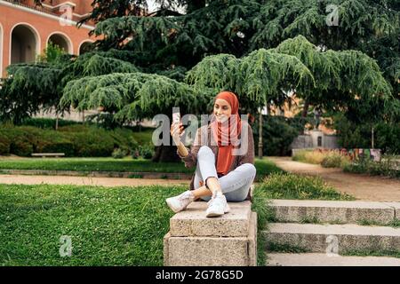 Bella donna musulmana che indossa hijab seduto nel parco e prendendo un selfie con il suo telefono. Foto Stock