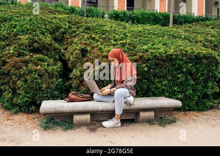 Bella giovane donna che indossa hijab seduto nel parco e utilizzando il suo computer portatile. Foto Stock
