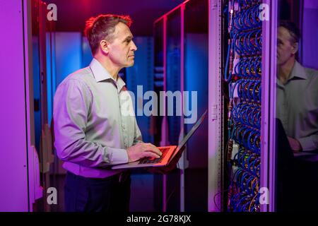 Concentrato maturo tecnico di rete caucasico in camicia in piedi presso un armadio server aperto ed eseguire il controllo di sicurezza utilizzando un laptop Foto Stock