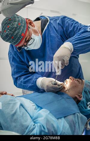 Dentista maschile indossando maschera facciale e indumenti protettivi che lavorano in clinica dentale con un paziente. Foto Stock