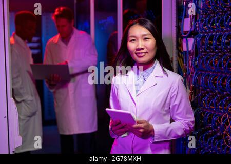 Ritratto di sorridente giovane ingegnere di rete asiatico femminile in camice bianco utilizzando tablet digitale contro cabinet con cavi Foto Stock