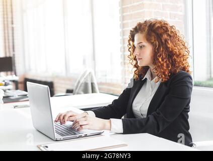Bella giovane donna con capelli rossi in un vestito da lavoro lavora in un ufficio con un computer portatile Foto Stock