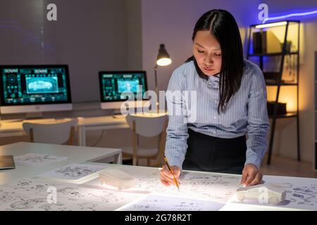 Concentrato giovane donna asiatica designer di automobili in piedi alla scrivania e disegno di disegni di auto esterno in ufficio scuro Foto Stock