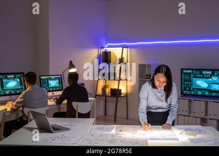 Giovane donna asiatica impegnata in camicia in piedi al tavolo con blueprint e disegno con matita, mentre lo sviluppo di auto design in ufficio Foto Stock