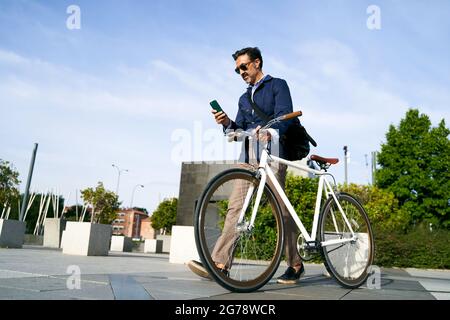 Uomo adulto che cammina in ufficio con una mano che tiene la bicicletta e l'altra mano che guarda il suo telefono cellulare. Concetto di trasporto ecocompatibile. Foto di alta qualità Foto Stock