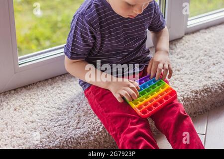 Un piccolo ragazzo biondo che gioca con un moderno colorato e luminoso popit giocattolo semplice concavità. Il bambino con colorati sili antistress alla moda Foto Stock