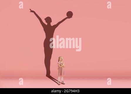 Infanzia e sogno sul grande e famoso futuro. Immagine concettuale con la ragazza e l'ombra di fit femminile ritmo ginnastica su sfondo corallo rosa Foto Stock