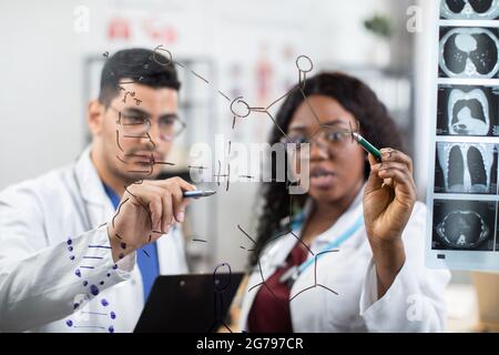Vista attraverso una parete di vetro. Offuscare il team creativo di due medici scienziati multirazziali, scrivendo sulla parete e guardando le formule chimiche. Medicina, concetto di biochimica Foto Stock