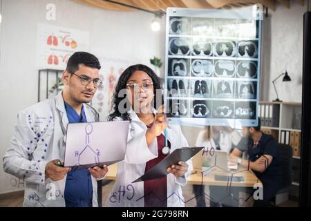 Gruppo di due medici multirazziali, in piedi dietro la parete di vetro, utilizzando un computer portatile durante l'esame della tomografia del paziente durante il consilium. Collegiesinternazionali che discutono varie modalità di trattamento. Foto Stock