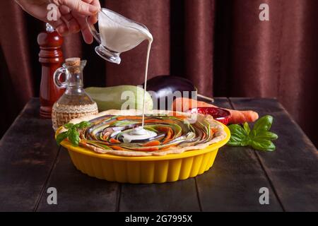 Processo di cottura della crostata a spirale vegetale con zucchine, melanzane, carote, peperone, basilico in teglia siliconica. La donna versa la crema su una torta. Foto Stock