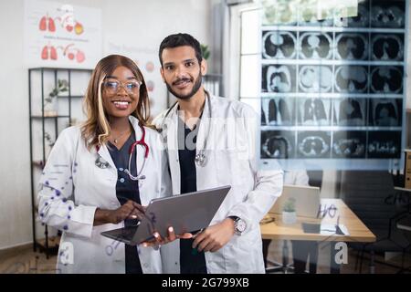 Vista attraverso la parete di vetro. Piacevole team di medici multietnici scienziati, addetta afro-americana e uomo arabo, in piedi in un ufficio luminoso, lavorando su un computer portatile e sorridendo alla telecamera Foto Stock