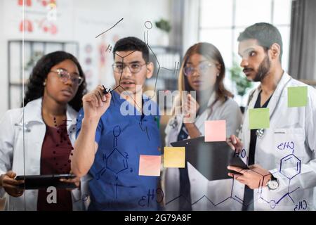 Gruppo di quattro team multietnici di medici, che discutono di fronte alla parete di vetro, scrivendo formule chimiche, utilizzando note e adesivi presso un moderno ufficio luminoso in clinica. Vista attraverso il vetro Foto Stock