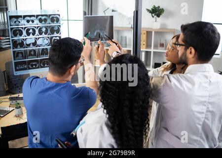 Gruppo di quattro medici multirazziali che cercano e discutono di raggi X in clinica o ospedale moderno. Team di medici multietnici che esaminano i raggi X della testa. Vista dall'angolo superiore Foto Stock