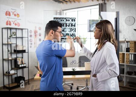 Donna africana e uomo indiano, medici concentrati in ospedale che guardano la scansione tc sulla parete di vetro. Medici ospedalieri di sesso femminile e maschile che controllano attentamente la scansione rm del paziente, esame regolare Foto Stock