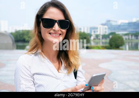 Ritratto di allegra donna di affari in occhiali da sole che risponde messaggi di testo sul suo smartphone Foto Stock