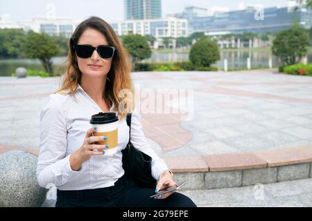 Ritratto di elegante donna d'affari con smartphone che riposa all'aperto, bevendo caffè da togliere e guardando la macchina fotografica Foto Stock