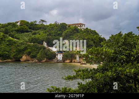 La vista da Ponte da Boa Viagem, Nitreói, Stato di Rio de Janeiro, Brasile. Isola verde Ilha da boa Viagem isola privata in baia collegata alla terra principale attraverso un ponte stretto e sabbia gialla sotto il paesaggio blu nuvoloso. Montagne in lontananza. Foto Stock