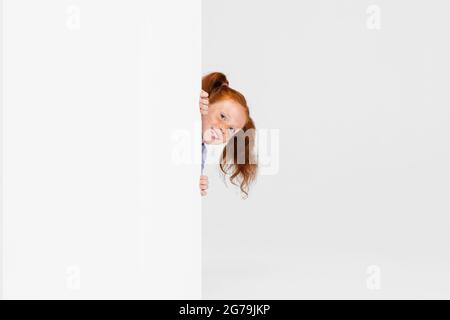 Ritratto di carina ragazza con testa rossa in abito casual sbucking fuori la parete isolato su sfondo bianco studio. Concetto di infanzia felice. Soleggiata Foto Stock