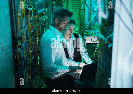 Visualizzazione ad angolo elevato presso due tecnici di rete che utilizzano un computer portatile nella sala server mentre lavorano con il supercomputer nel data center Foto Stock