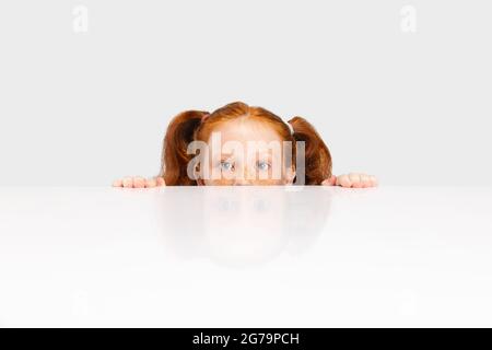 Ritratto di carina ragazza con testa rossa in abito casual sbucking fuori la parete isolato su sfondo bianco studio. Concetto di infanzia felice. Soleggiata Foto Stock