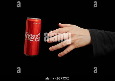 KALININGRAD, RUSSIA - 13 MARZO 2021 - la mano raggiunge la lattina di Coca Cola, sfondo nero. Classico vaso di coke, bevanda analcolica gassata. Prodotto dalla C Foto Stock