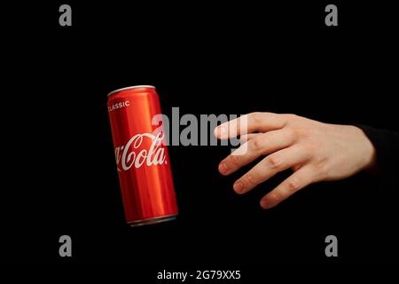 KALININGRAD, RUSSIA - 13 MARZO 2021 - la mano raggiunge la lattina di Coca Cola, sfondo nero. Classico vaso di coke, bevanda analcolica gassata. Prodotto dalla C Foto Stock