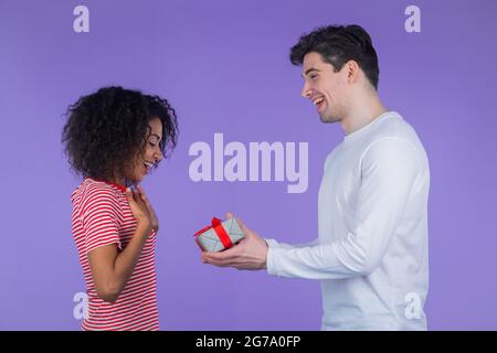 Giovane coppia interracial su sfondo studio viola. L'uomo bianco dà dono con arco rosso alla sua moglie o fidanzata afroamericana. Compleanno, amore Foto Stock
