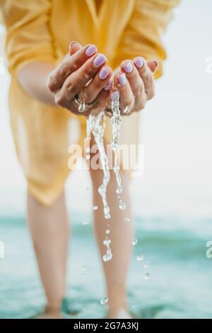 L'acqua che versa dalle mani del donna. Mare, vicino alle braccia femminili. Camicia gialla sullo sfondo. L'acqua è una forza vitale Foto Stock