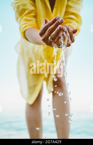 L'acqua che versa dalle mani del donna. Mare, vicino alle braccia femminili. Camicia gialla sullo sfondo. L'acqua è una forza vitale Foto Stock