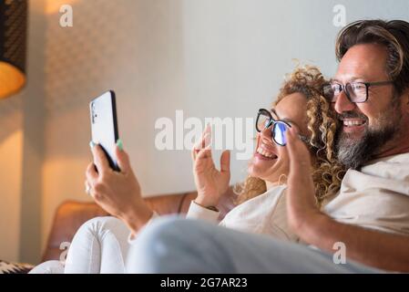 Happy Young Adult Couple Making Video Call on Phone - le persone allegre parlano e ascoltano gli amici online con il cellulare da casa seduta sul divano - festeggiamenti a distanza concetto Foto Stock