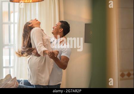 Felice etnia mista di razza marito e moglie si sentono eccitati ballare ondeggiando in piedi a casa, sorridendo coppia sopravorato divertirsi festeggiando anniversario swirling e ballare insieme in amore Foto Stock
